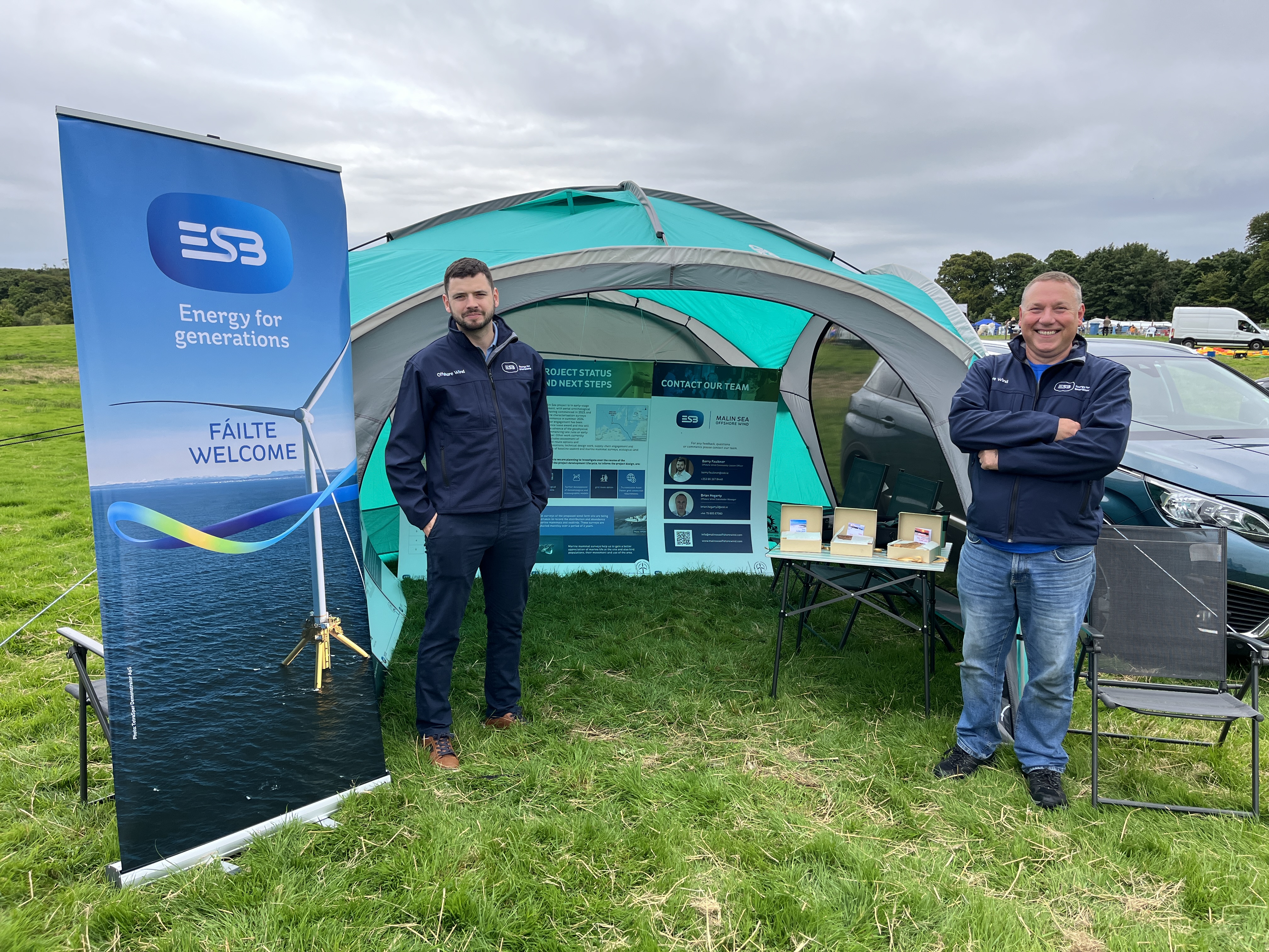 Two men standing at both sides of a camping tent with branded pop-up stand and camping chair and cars and vans in the background.